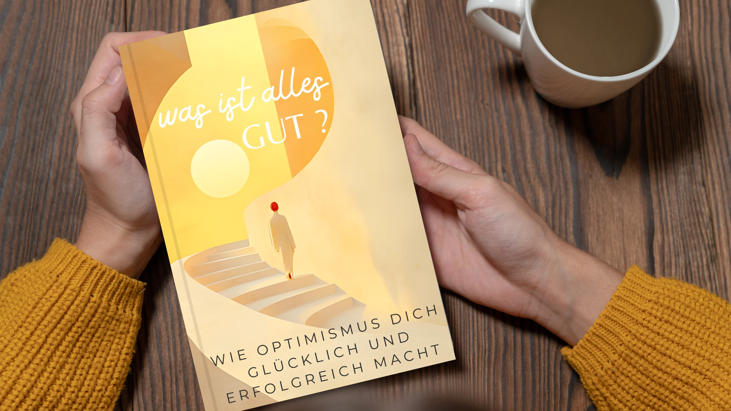 Ein Buchcover mit dem Titel "Was ist alles gut?", einem Text über Optimismus, gehalten in Händen, auf Holztisch mit Tasse daneben.